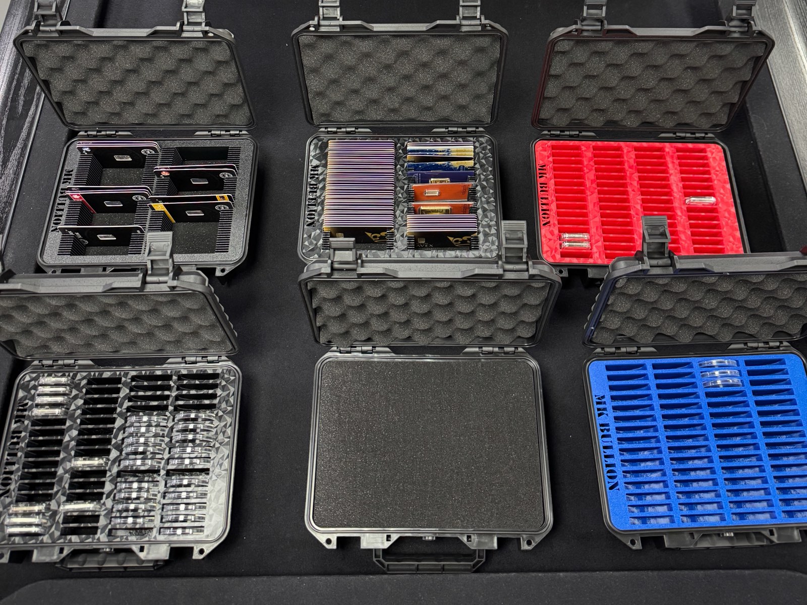 Six open MK Bullion cases showing different 3D-printed inserts — card bars, coin capsules, and red and blue bar trays